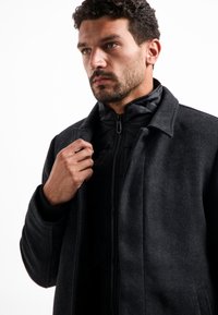 Kolenwollen gemêleerde jas met een textuur oppervlak, een kraag en een zwart met rits gesloten binnenlaag. Voorzien van een relaxte pasvorm en zijzakken.