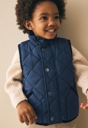 Usmievavé dieťa so zvlnenými vlasmi, oblečené v prešívanej námorníckej vestičke cez krémový pletený sveter, stojace na béžovom pozadí.