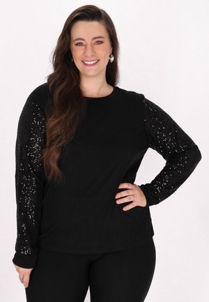 Sort langærmet skjorte med pailletbelagte ærmer, glat kropstof, rund halsudskæring og tætsiddende design. Båret med sorte bukser.