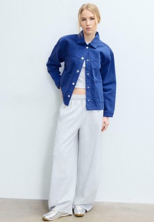 Młoda kobieta ubrana w niebieską kurtkę na guziki, biały krótki top, szerokie jasnoszare spodnie dresowe i srebrne tenisówki, stoi na tle gładkiej ściany.