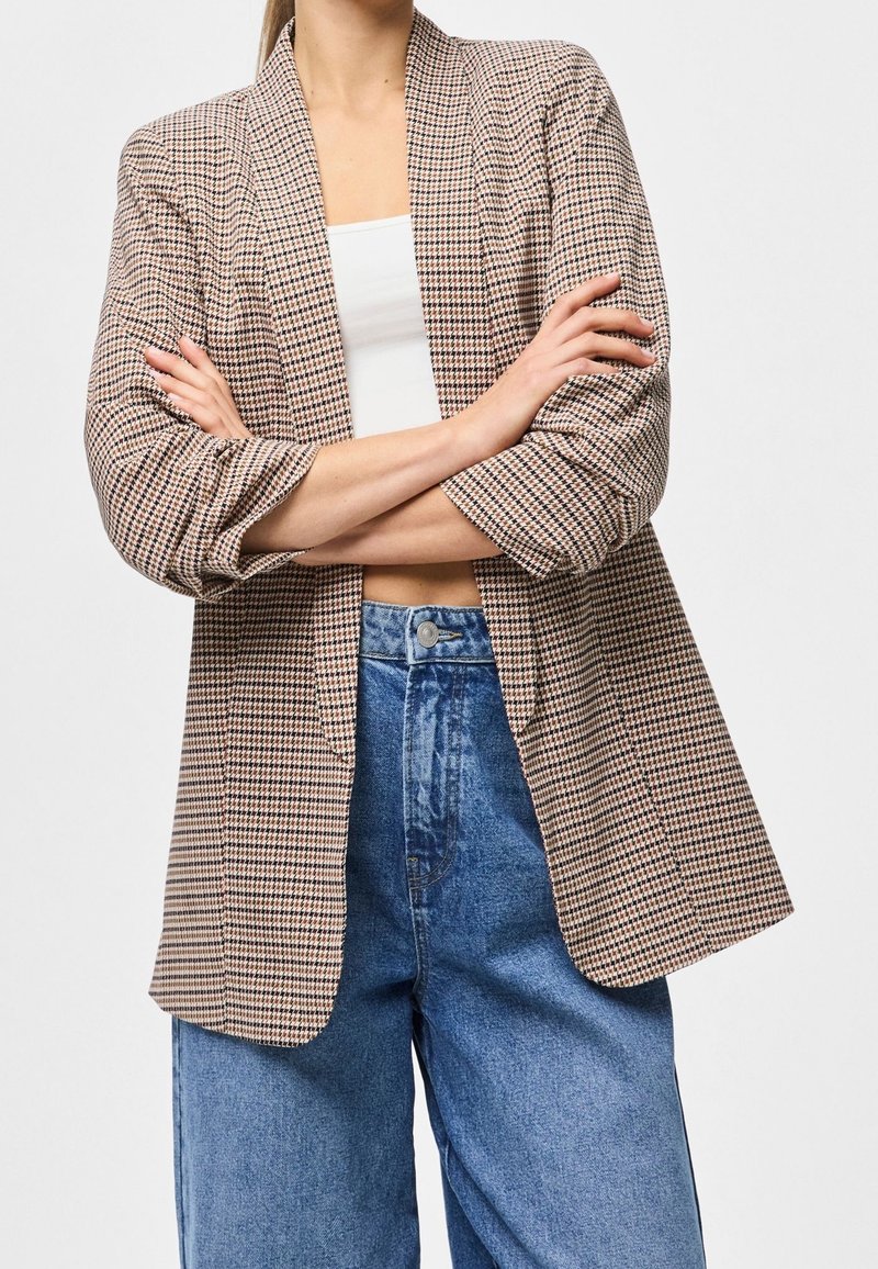 Personne portant un blazer à carreaux sur un crop top blanc et un jean bleu taille haute, bras croisés devant le corps.