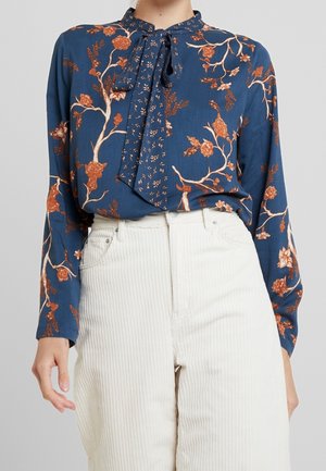 Niebieska bluzka z długim rękawem z pomarańczowym kwiatowym nadrukiem i zawiązanym szalikiem na szyi, zestawiona z białymi, spodniami sztruksowymi z wysokim stanem.