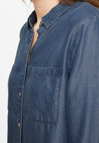 Denim shirt in dark blue, featuring a button-down collar, two chest pockets, and a soft texture. Buttons are a shiny metallic color.
