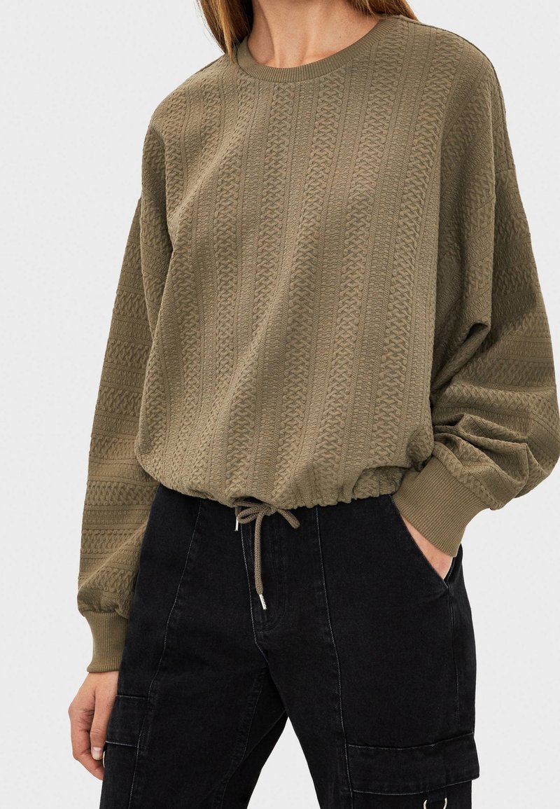 Femme portant un pull en maille texturée vert olive avec taille à cordon de serrage et pantalons noirs taille haute avec poches, une main dans la poche.