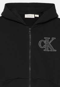 Crna dukserica s patentnim zatvaračem, od mekog materijala, s logotipom "ck" u srebrnoj sjaju na lijevoj strani prsnog koša i kapuljačom s utezima.