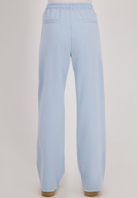 Lichtblauwe, losse broek met elastische tailleband en twee achterzakken, gedragen met beige schoenen op een neutrale achtergrond.