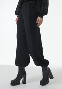 Zwarte broek heeft gestructureerde zijpanelen, taps toelopende boorden en een ontspannen pasvorm. Gecombineerd met zwarte hoge hakken. Het materiaal is glad.