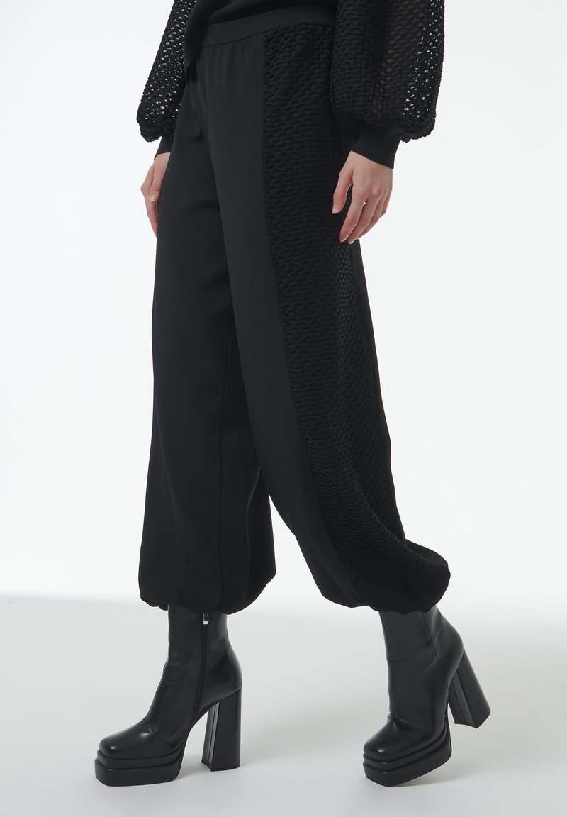 Zwarte broek heeft gestructureerde zijpanelen, taps toelopende boorden en een ontspannen pasvorm. Gecombineerd met zwarte hoge hakken. Het materiaal is glad.