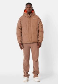 Bruine pufferjas met een ritssluiting aan de voorkant, capuchon en oranje voering, gedragen over bijpassende bruine broeken en witte sneakers. Zachte textuur, vierkante vorm.