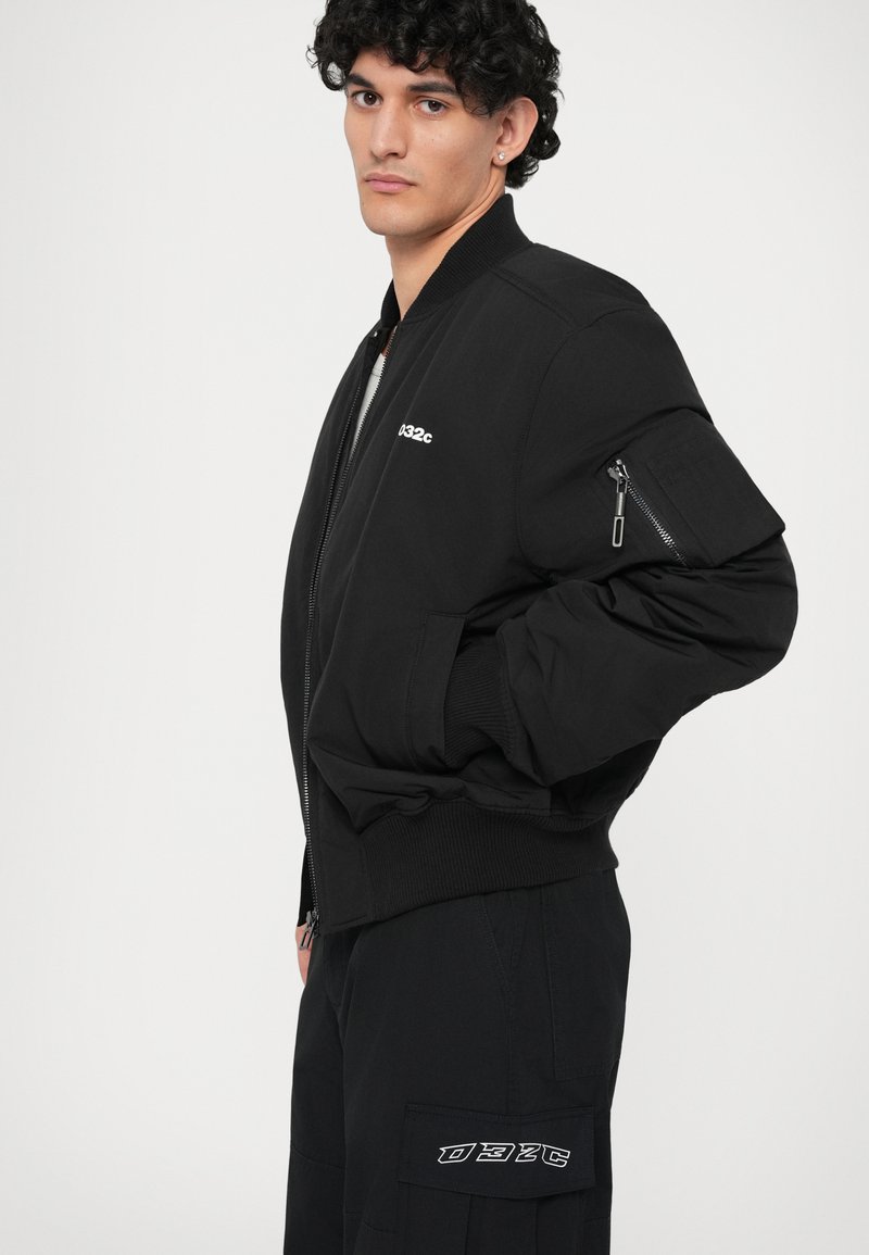 Schwarze Bomberjacke mit gerippten Bündchen und Kragen. Mit einer Reißverschlusstasche am Ärmel und einem Logopatch. Glatte Stoffoberfläche.