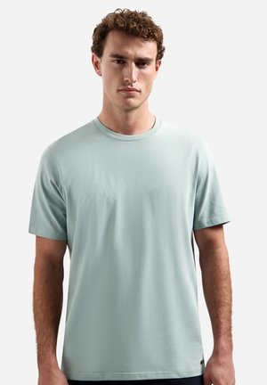 Junger Mann mit lockigem Haar, der ein hellblaues kurzärmliges Rundhals-T-Shirt trägt, steht vor einem schlichten weißen Hintergrund.