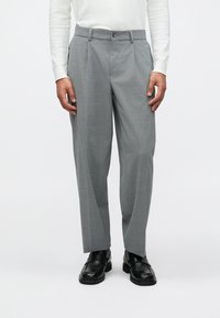 Calças cinzas feitas à medida com uma textura subtil, apresentando um corte reto, bolsos inclinados e um fecho frontal com botão. Usadas com sapatos pretos.