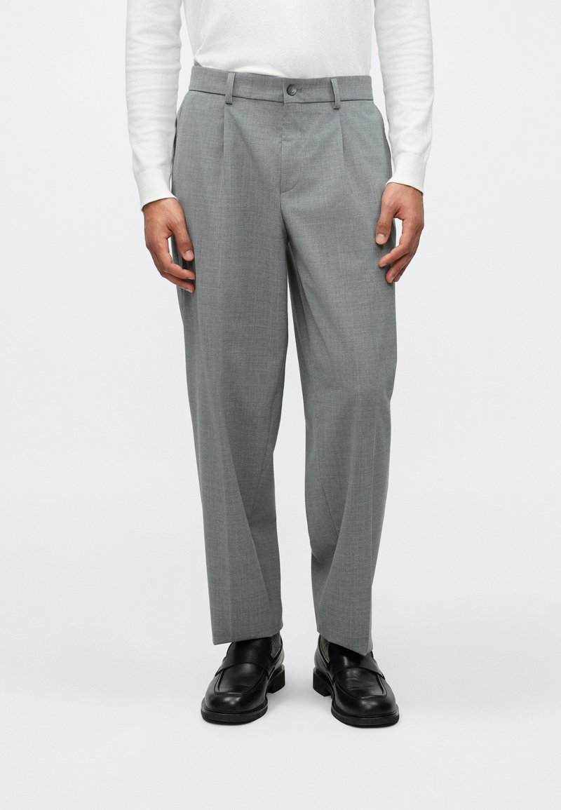 Calças cinzas feitas à medida com uma textura subtil, apresentando um corte reto, bolsos inclinados e um fecho frontal com botão. Usadas com sapatos pretos.