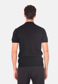 Un uomo sta in piedi di spalle indossando una maglietta nera a maniche corte e pantaloni scuri su uno sfondo chiaro e neutro.