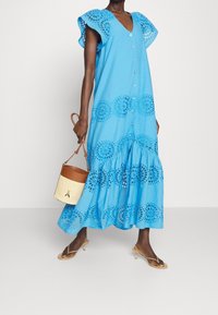 Robe longue en coton bleu clair avec détails en œillets torsadés, col en V et boutons sur le devant. Associée à un sac seau beige et des sandales à lanières.