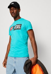 Turquoise short-sleeve t-shirt with a black Diesel logo on the chest. Model holds a black jacket with orange lining. Casual fit.