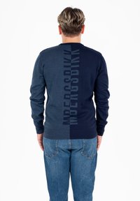 Mężczyzna z krótkimi blond włosami ubrany w niebieski sweter w dwóch odcieniach z pionowym napisem i niebieskie dżinsy, stojący tyłem na białym tle.