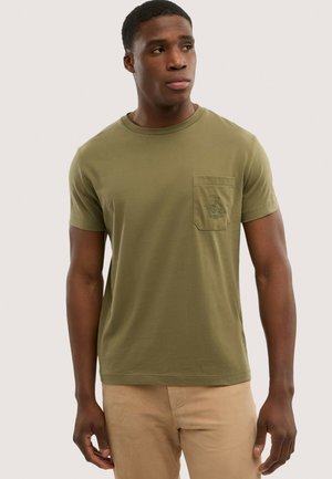 Giovane uomo che indossa una t-shirt girocollo verde oliva con una tasca sul petto e pantaloni beige, che guarda verso destra.