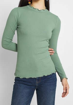 Femme portant un haut côtelé vert clair à manches longues avec des bords à volants et un jean bleu foncé, posant avec une main sur la hanche.