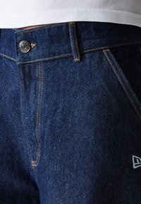 Jeans en denim bleu foncé avec des coutures orange contrastantes, fermeture à bouton en métal et un petit logo blanc sur la jambe inférieure.