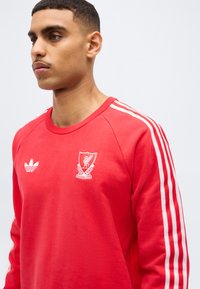 Noor mees, kes kannab punast Liverpooli jalgpalliklubi dressipluusi valgete Adidas triipudega varrukatel, vaadates vasakule ühevärvilise tausta ees.