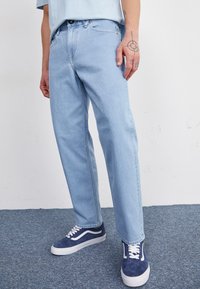 Jeans en denim bleu clair à coupe droite, présentant un design classique à cinq poches et des détails de couture. Portés avec des baskets bleu marine.