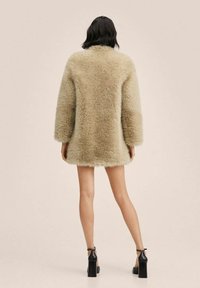 Femme vue de dos portant un manteau en fausse fourrure beige et des chaussures noires à talons hauts avec bride à la cheville, debout devant un fond uni.