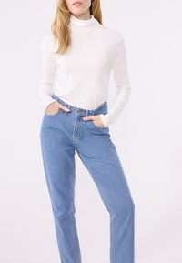 Eine Frau trägt einen weißen Rollkragenpullover und blaue hochgeschnittene Jeans, die Hände in den Taschen, und steht vor einem schlichten hellen Hintergrund.