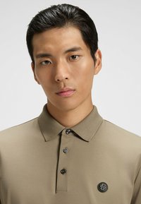 Khaki polo z złożonym kołnierzykiem, z zapięciem na dwa guziki i małą czarną łatką z logo na piersi. Gładka faktura i dopasowany krój.