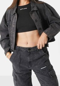 Donna indossa un top corto a coste neri con la scritta "SIXTH JUNE", una giacca in denim nera lavata e pantaloni cargo abbinati con tasche.