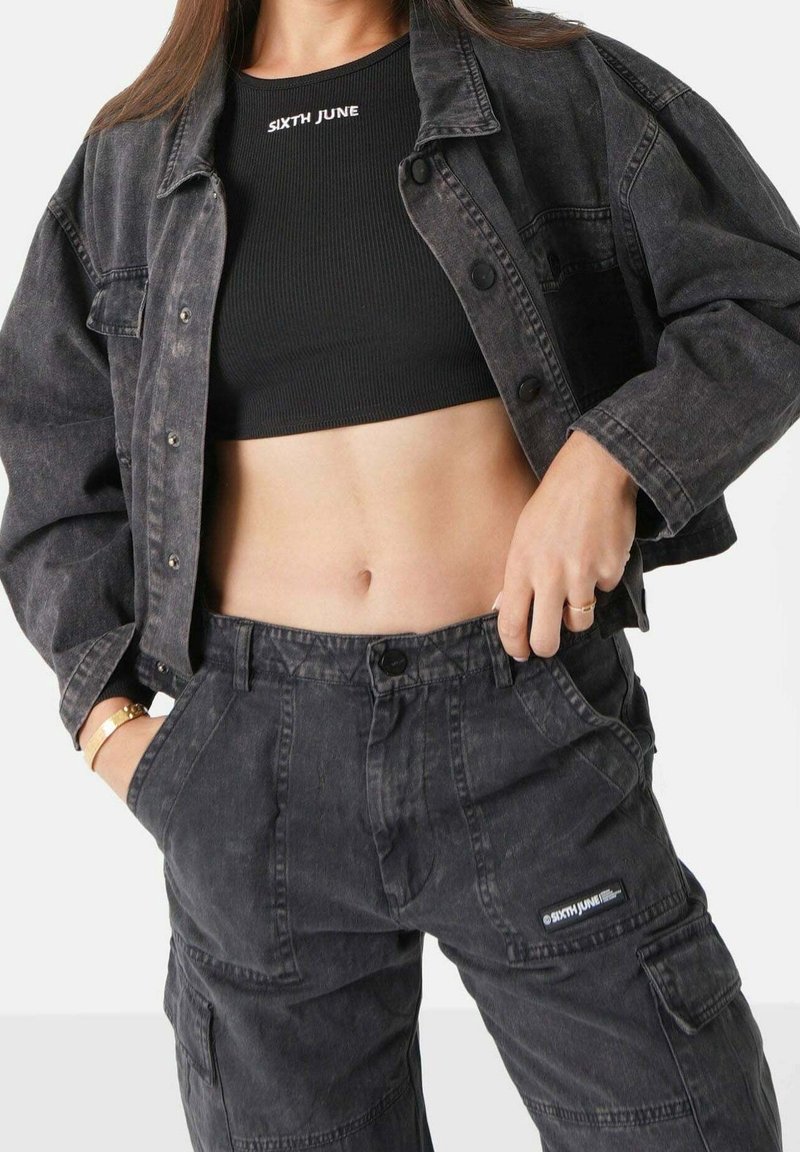 Donna indossa un top corto a coste neri con la scritta "SIXTH JUNE", una giacca in denim nera lavata e pantaloni cargo abbinati con tasche.