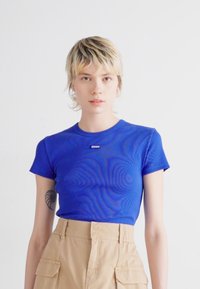 Maglietta aderente blu con maniche corte e scollatura rotonda, caratterizzata da un piccolo etichetta con logo sul davanti. Colore uniforme, texture liscia.