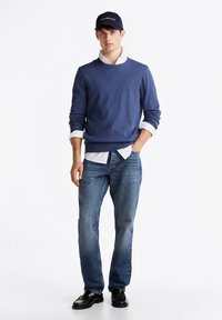 Camisola azul com decote redondo, usada sobre uma camisa branca, combinada com jeans azul claro e sapatos pretos. Acessorizada com um boné escuro.