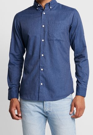 Mann trägt ein blaues, langärmeliges Hemd mit Knöpfen und Brusttasche sowie hellblaue Jeans, steht vor einem einfarbigen Hintergrund.