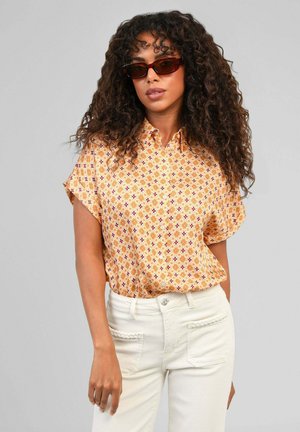 Femme aux cheveux bouclés portant des lunettes de soleil marron, une chemise jaune à fleurs à manches courtes boutonnée et un pantalon blanc sur fond gris.
