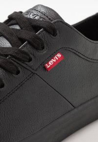 Svart lädersneaker med texturerad yta, runda svarta snören, svarta ögla och en röd Levi's-logotyp på sidan. Enkel design.