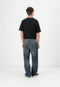 Camiseta de algodón negra combinada con jeans de denim azul desgastados de corte holgado, que cuentan con dos bolsillos traseros y una etiqueta de cuero en la cintura. Zapatillas negras y blancas.