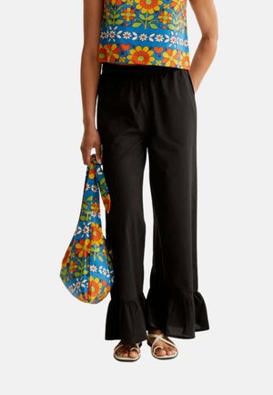 Persona que lleva pantalones negros de pierna ancha con dobladillos con volantes, sandalias blancas y sostiene una bolsa azul con estampado floral que combina con su parte superior.