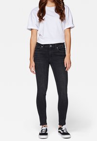 Pantalones vaqueros ajustados de lavado oscuro con cintura media, que cuentan con bolsillos frontales, cierre de botón y detalles de costura sutiles, combinados con una camiseta blanca.