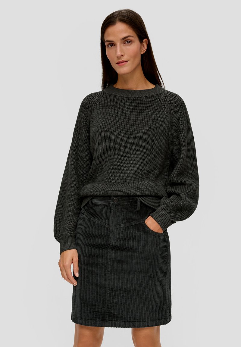 Schwarzer gerippter Strickpullover mit hohem Kragen und langen Ärmeln, kombiniert mit einem knielangen schwarzen Cordrock mit Vordertaschen.