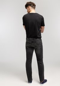 Mężczyzna stojący z założonymi rękami, ubrany w czarną koszulkę, ciemnoszare dżinsy i granatowe trampki na jednolitym szarym tle.