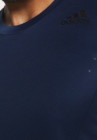 Detailní záběr osoby v tmavě modrém sportovním tričku Adidas se strukturou, na kterém se nachází logo na horní části hrudníku blízko ramene.