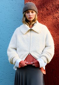 Krémová fleecová bunda se širokým límcem a předním zipem, spárovaná s rustikově zbarveným vrstveným topem a šedou sukní, na pozadí s texturou.