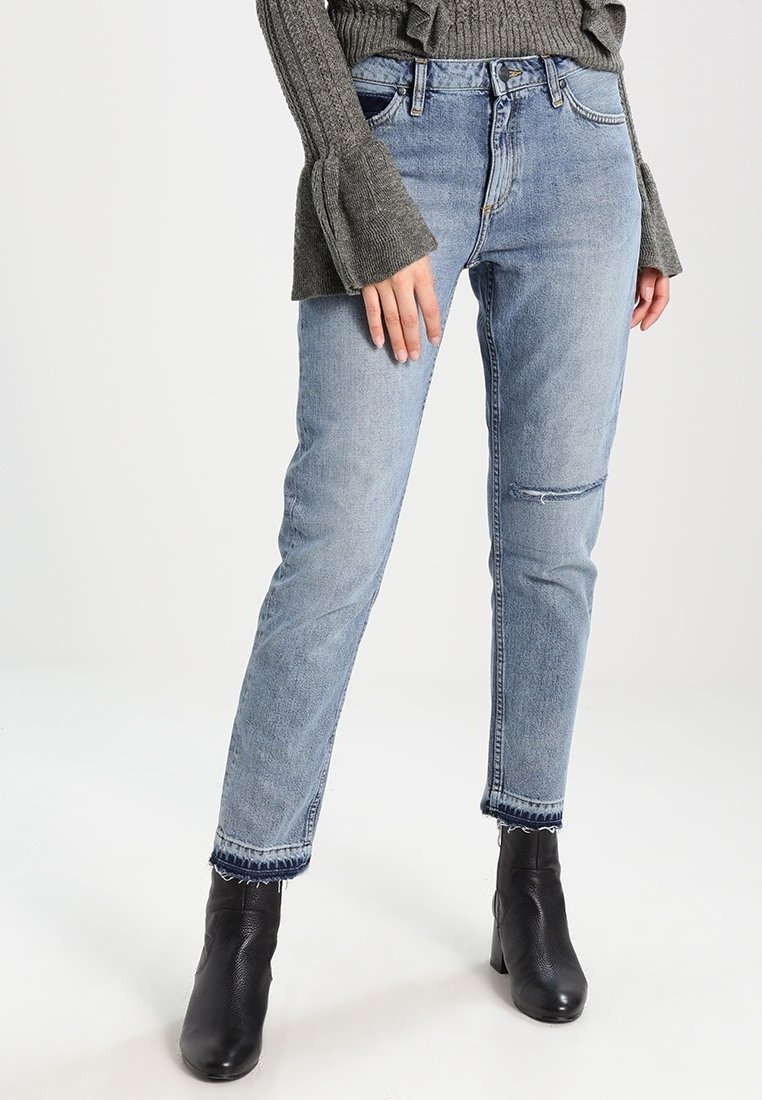 Jean en denim bleu clair coupe décontractée, avec des détails déchirés au niveau des genoux et des ourlets effilochés, assorti à des bottines noires.