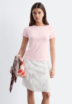 Jeune femme portant un haut rose clair à manches courtes et une jupe blanche à fleurs, tenant une écharpe à motifs dans sa main gauche sur un fond uni.