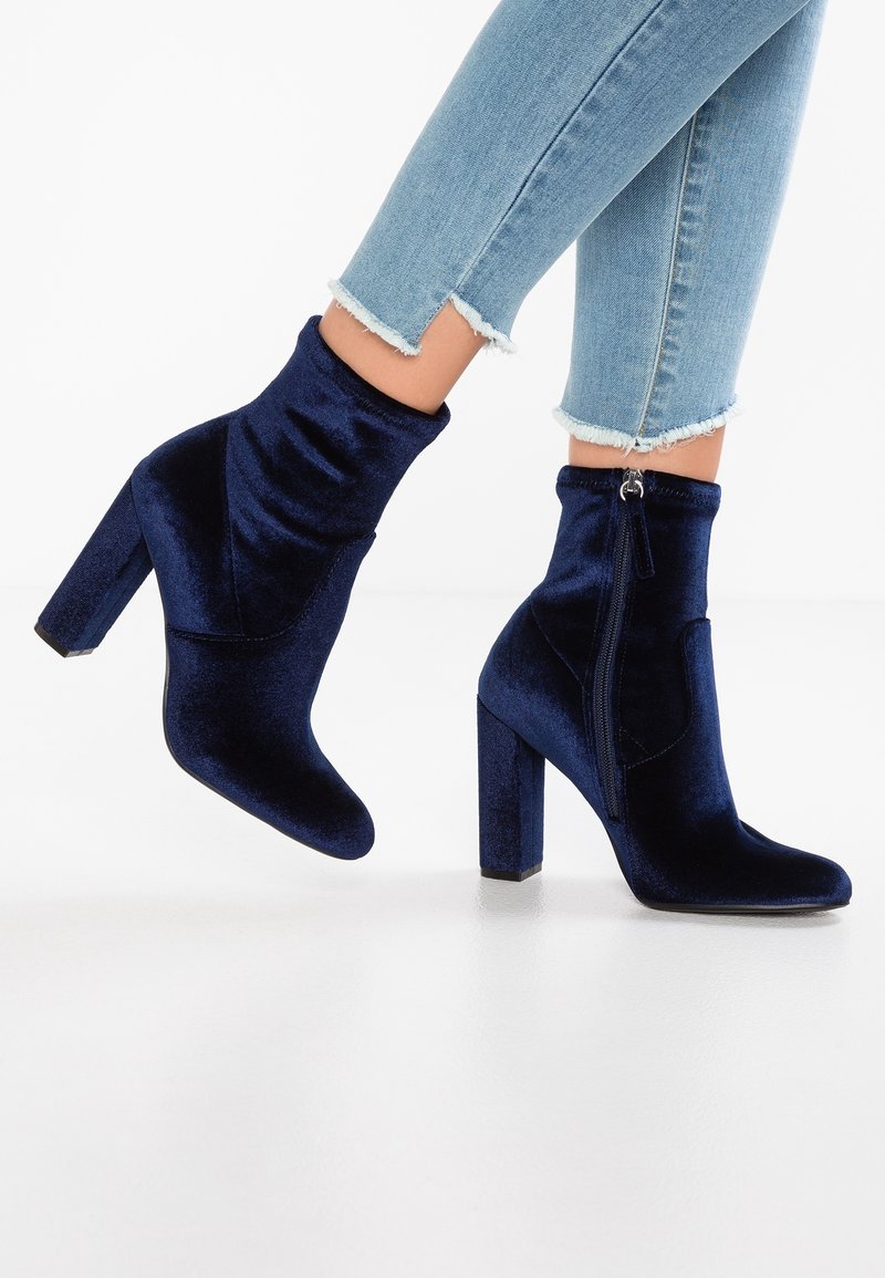 Femme portant un jean skinny bleu clair et des bottines en velours bleu marine avec talons blocs et fermetures éclair latérales.
