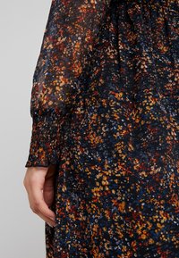 Květinové šaty s tmavým základem a barevnými květy v červené, žluté a modré barvě. Průhledná látka, smockované manžety a volný střih.