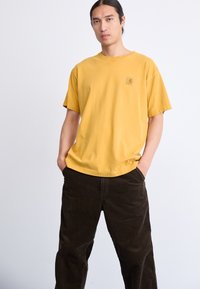Gul bomulds T-shirt med rund hals og korte ærmer, der har et lille logo patch på venstre bryst. Bæres med mørkebrune fløjlskomfiner.