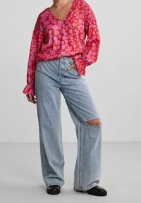 Blusa a pois rossi e rosa con scollatura a V e maniche a sbuffo, abbinata a jeans a gamba larga strappati di colore blu chiaro e scarpe nere.