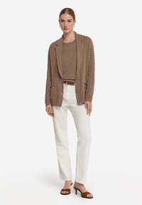 Brun stickad cardigan med en sjalkrage, framfickor och knappar, i kombination med en brun topp och vita byxor, kompletterad med bruna klacksandaler.